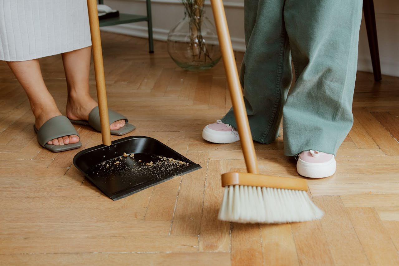 Close-up of people cleaning wooden floor with broom and dustpan indoors.