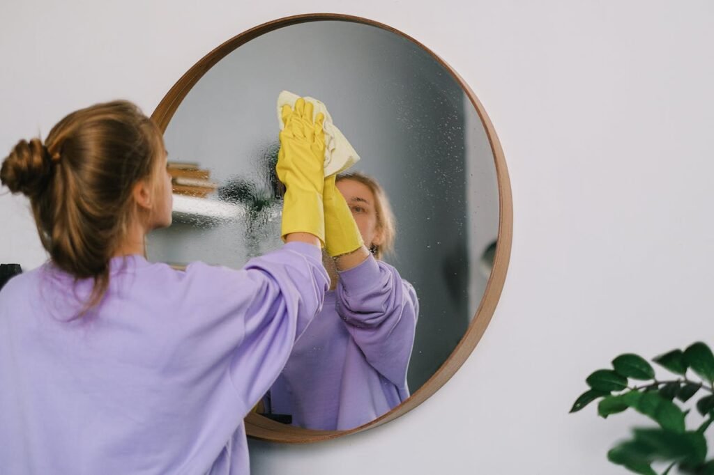 A woman wearing yellow gloves cleans a round wall mirror indoors, reflecting her image.